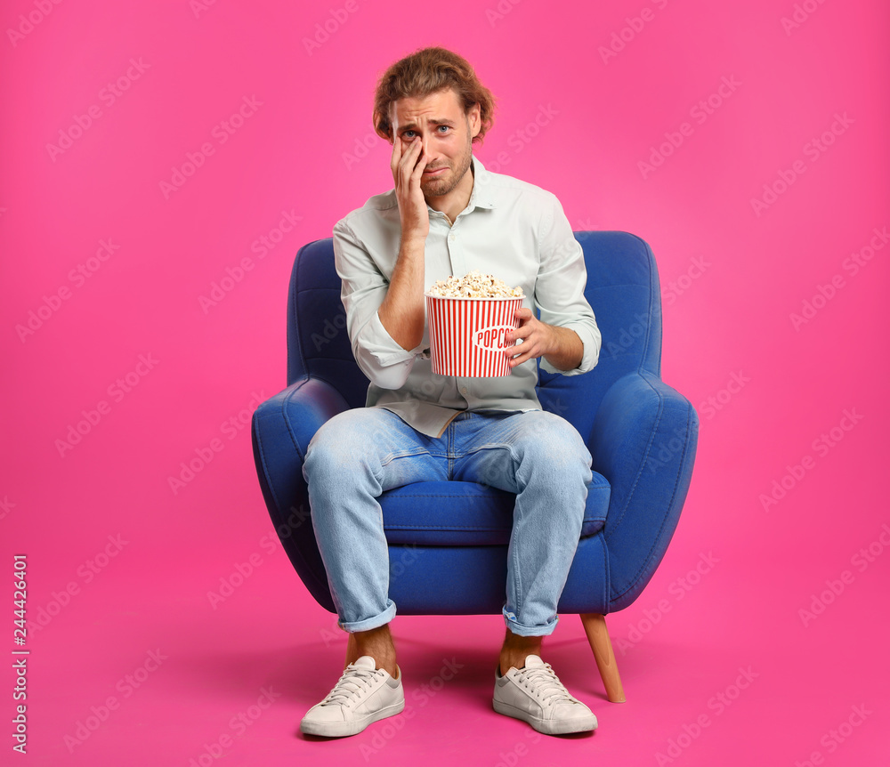 Fototapeta premium Emotional man with popcorn sitting in armchair during cinema show on color background
