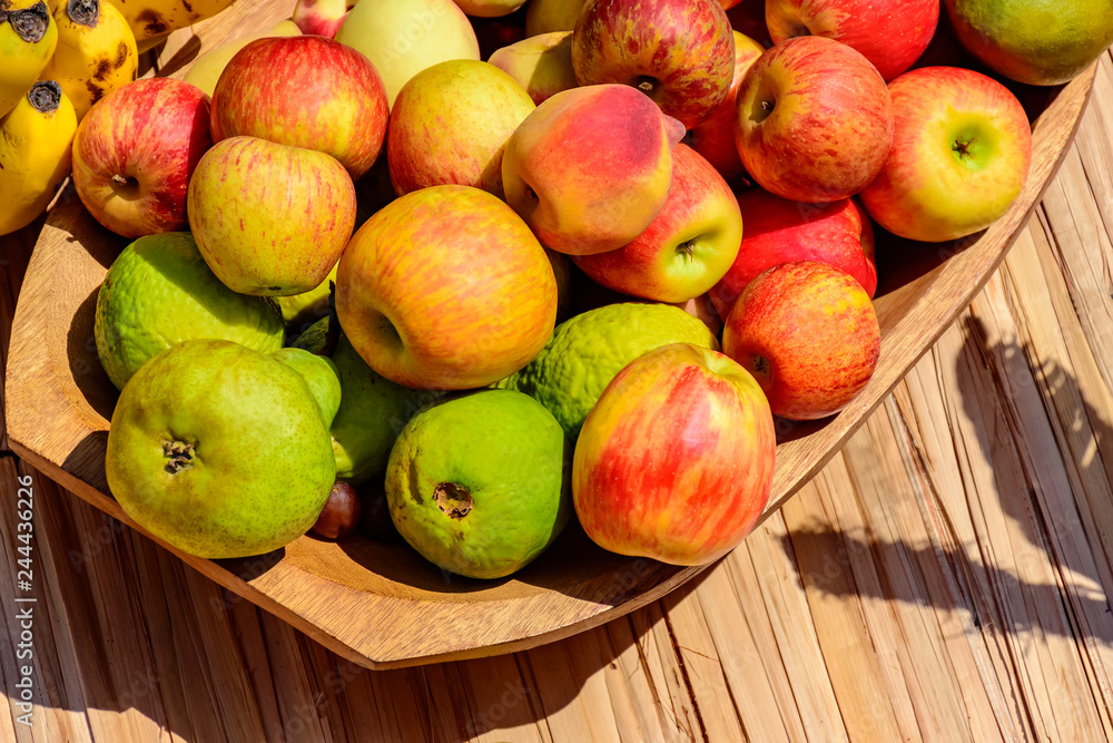 Wooden bowl and rustic straw mat with various guavas, apples and bananas under the sunlight
