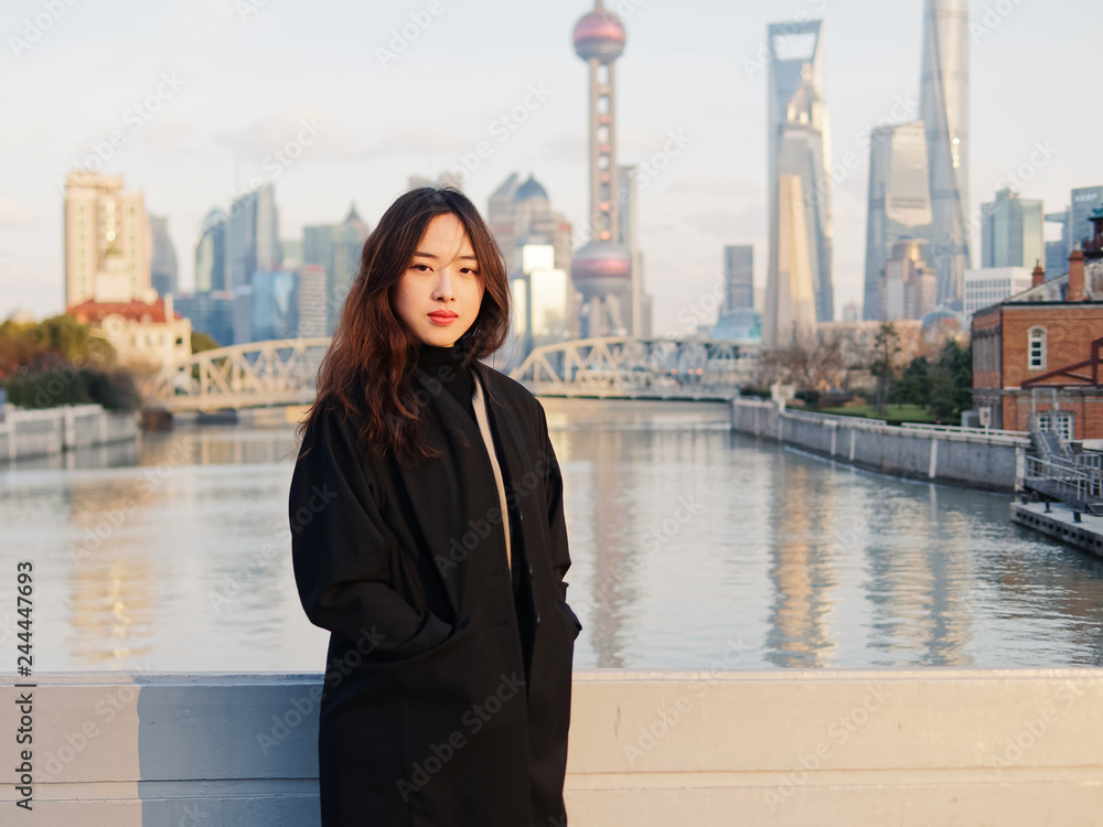 Fototapeta premium Beautiful young brunette woman in black clothes smiling at camera with blur Shanghai Bund landmark buildings background with dusk light. Emotions, people, beauty, travel and lifestyle concept.