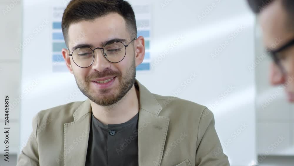 Tracking close up shot of friendly bearded young businessman in glasses smiling while talking to group of colleagues at meeting Tracking close up shot of friendly bearded young businessman in glasses smiling while talking to group of colleagues at meeting