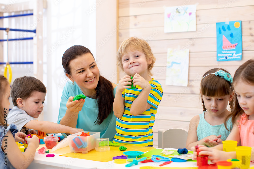 Fototapeta premium Children dough play in daycare centre. Kids with teacher mold from plasticine in kindergarten. Little students knead modeling clay with hands in preschool.
