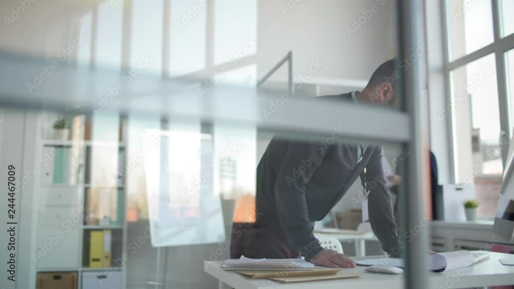 Tracking right of Caucasian office worker coming to his workplace and starting using computer while his male coworker standing by window with documents in his hands Tracking right of Caucasian office worker coming to his workplace and starting using computer while his male coworker standing by window with documents in his hands