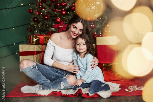 Holidays conception. Cheerful mother and daughter sitting near the Christmas tree that behind. Cute portrait © standret