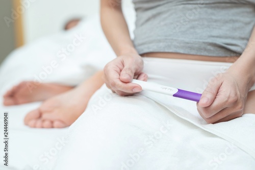 Young woman looking show in hand at checking pregnancy test kit. She's is checking her pregnancy exam sitting on the bed with couple lovers in bedroom. Premature is pregnant before age.