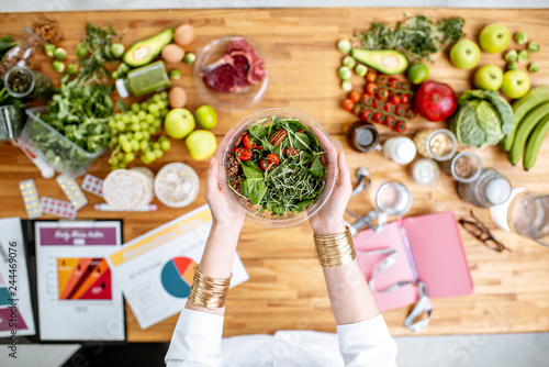 Dietitian holding cooked salad above the table full of various healthy products and drawings on the topic of healthy eating photo
