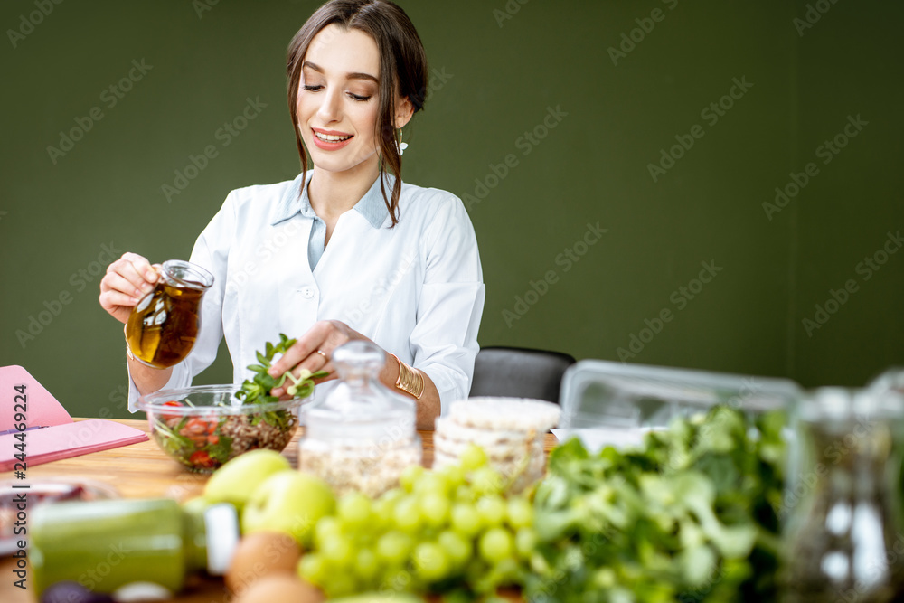 Woman dietitian working on a healthy diet pouring olive oil into the salad sitting at the table full of various healthy food indoors Woman dietitian working on a healthy diet pouring olive oil into the salad sitting at the table full of various healthy food indoors