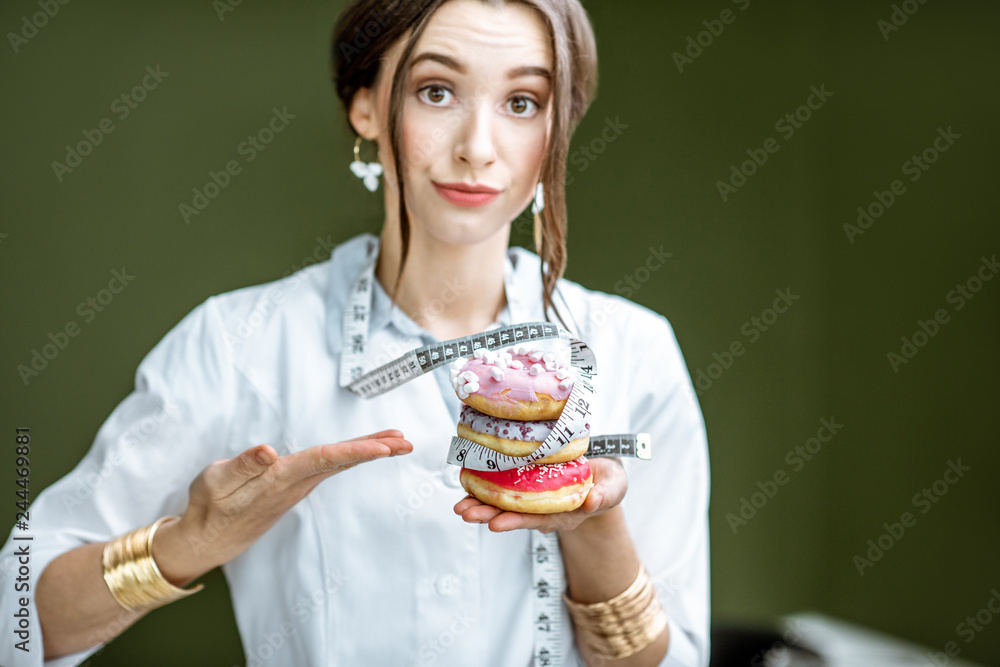 Portrait of a sad woman nutritionist in medical gown with donuts and tape measure on the green background. Unhealthy eating and adiposity concept Portrait of a sad woman nutritionist in medical gown with donuts and tape measure on the green background. Unhealthy eating and adiposity concept