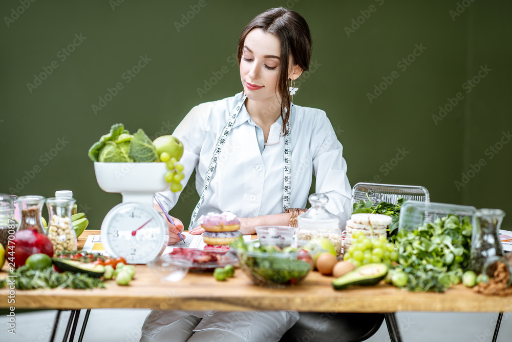 Woman dietitian in medical uniform working on a diet plan sitting with various healthy food ingredients in the green office Woman dietitian in medical uniform working on a diet plan sitting with various healthy food ingredients in the green office