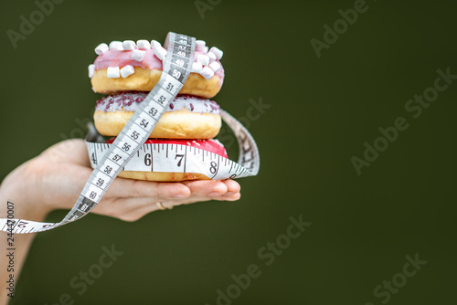 Three sweet donuts covered with measurement tape on the hand on the green background. Unhealthy eating and adiposity concept