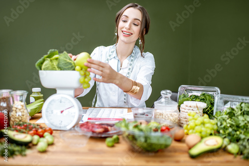 Woman dietitian in medical uniform working on a diet plan weighting fruits sitting with various healthy food ingredients indoors