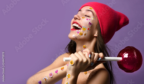 Brunette woman with popular social emoji smiles stickers on her face and hands happy smiling laughing hold big sweet lolly pop candy