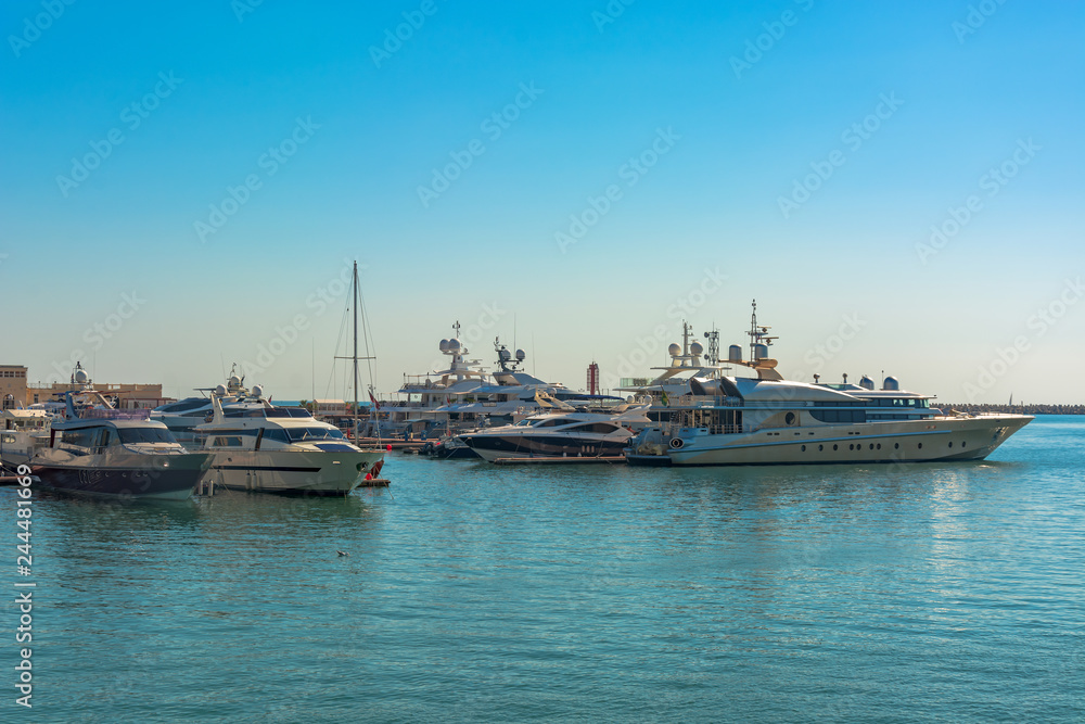 Sea port in summer. Many different yachts and boats stand at the marina. Yachtsmen and travelers are preparing to sail in the sea. In the background of the building of the seaside city. Sea port in summer. Many different yachts and boats stand at the marina. Yachtsmen and travelers are preparing to sail in the sea. In the background of the building of the seaside city.