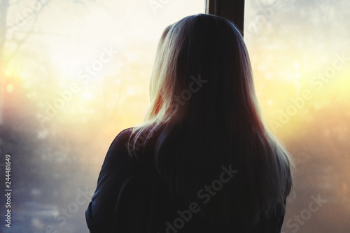 Blonde woman looking out the window into the morning light, back to the camera