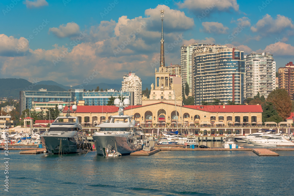 Fototapeta premium Sea port in summer. Many different yachts and boats stand at the marina. Yachtsmen and travelers are preparing to sail in the sea. In the background of the building of the seaside city.