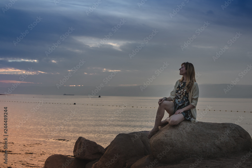 Fototapeta premium Girl on the rocks above sea and evening sky with sunset and blue clouds
