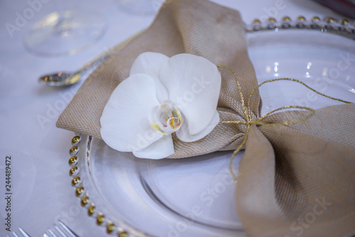 Fototapeta Naklejka Na Ścianę i Meble -  Fine dining table setting featuring transparent plates, beige linen napkin with natural orchid and golden decorations and silverware in the order of use, ready for guests at a formal event or wedding