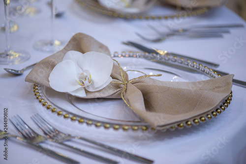 Fine dining table setting featuring transparent plates, beige linen napkin with natural orchid and golden decorations and silverware in the order of use, ready for guests at a formal event or wedding