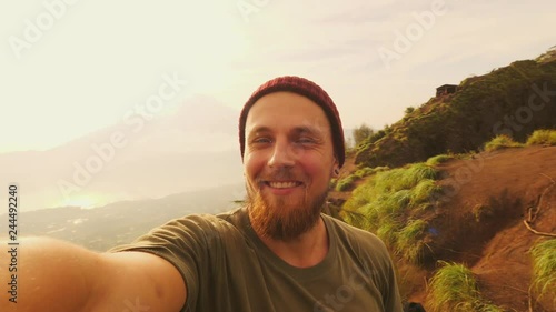 Young Caucasian Bearded Hipster Blogger Taking Selfie Portrait Video on Top of Batur Volcano with Beautiful Natural Sunrise Landscape on Background. 4K Slowmotion Retro Lifestyle Hiking Travel Concept