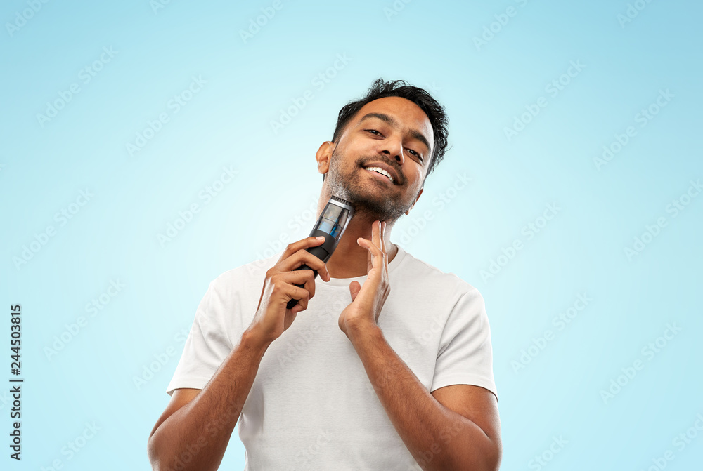 grooming, technology and people concept - smiling indian man shaving beard with trimmer over blue background grooming, technology and people concept - smiling indian man shaving beard with trimmer over blue background