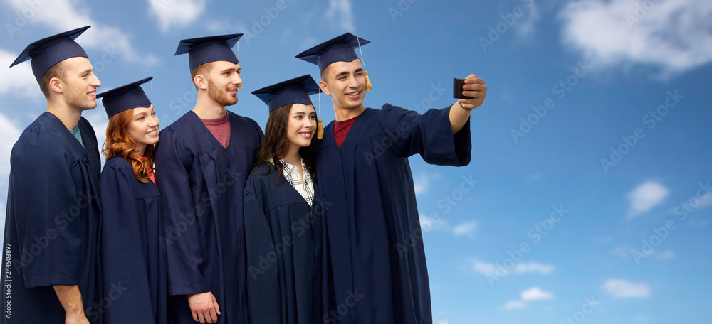 education, graduation and people concept - group of happy graduate students in mortar boards and bachelor gowns taking selfie by smartphone over blue sky and clouds background
