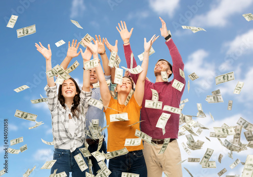 finance, wealth and people concept - group of happy friends picking money falling from up above over blue sky and clouds background