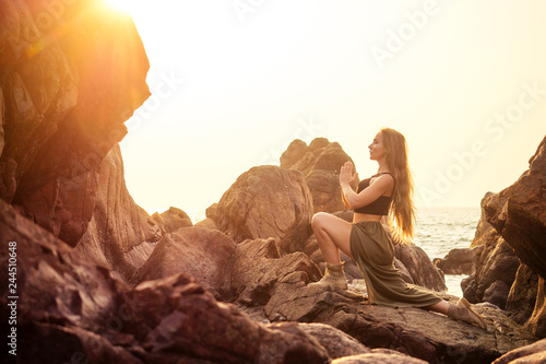 stylish redheaded chestnut blonde young girl in indie bohemian bo-ho style clothes black stylish top.boho woman long hair,makeup and green feathers in head practicing yoga asanas on the rocks by sea