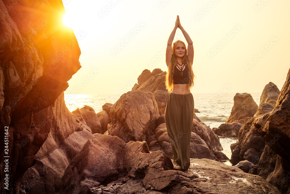 stylish redheaded chestnut blonde young girl in indie bohemian bo-ho style clothes black stylish top.boho woman long hair,makeup and green feathers in head practicing yoga asanas on the rocks by sea - obrazy, fototapety, plakaty 