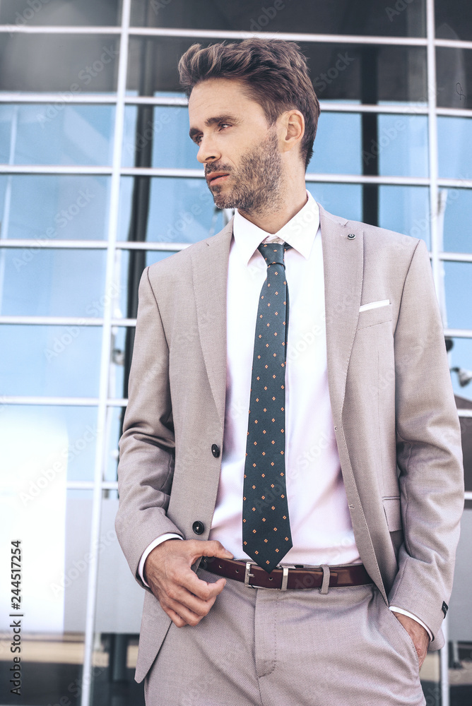 Young attractive serious businessman is standing next to a glassy office building wearing light classic tuxedo. Young attractive serious businessman is standing next to a glassy office building wearing light classic tuxedo.