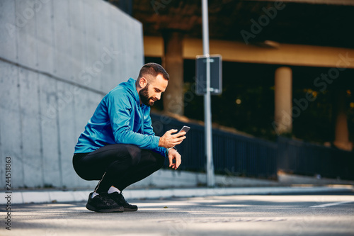 Young Caucasian bearded sporty man with short hair in sportswear crouching on the street and looking at smart phone. Healthy lifestyle concept.