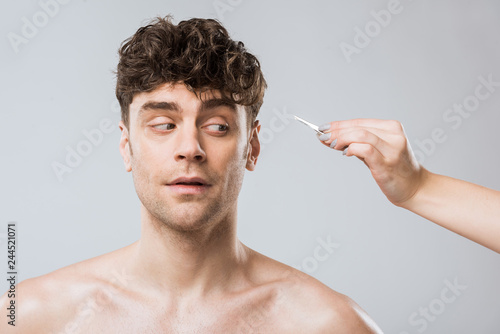 female hand plucking hair with tweezers from mans face  isolated on grey