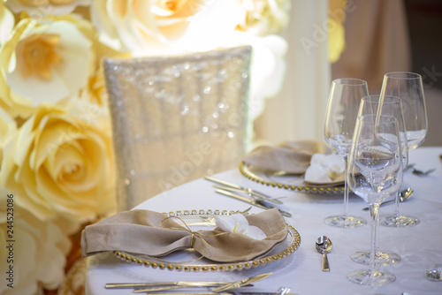 Sitting arrangement at a formal event or fine dining restaurant featuring transparent plates with golden details, glassware and silverware in the order of use set against a paper flowers wall