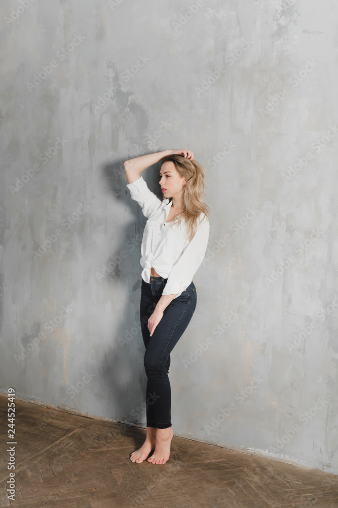 Model blonde blue jeans white shirt standing gray wall by window copy space. Model blonde blue jeans white shirt standing gray wall by window copy space.