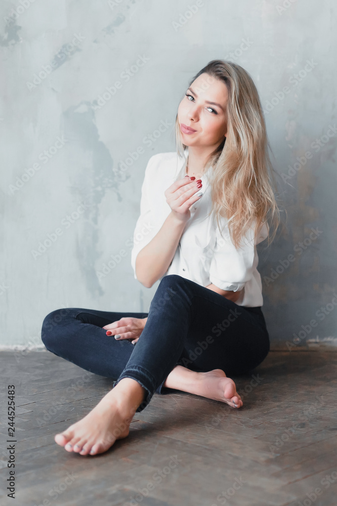 Model blonde barefoot blue jeans white shirt sitting on floor against gray wall.
