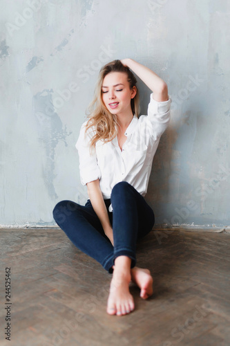 Model blonde barefoot blue jeans white shirt sitting on floor against gray wall.