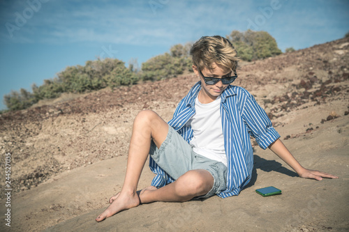 Blond child with sunglasses sitting on the beach looks at the cellphone. excessive use of the cellphone. smartphone generation. Blue sky and rocks in background photo