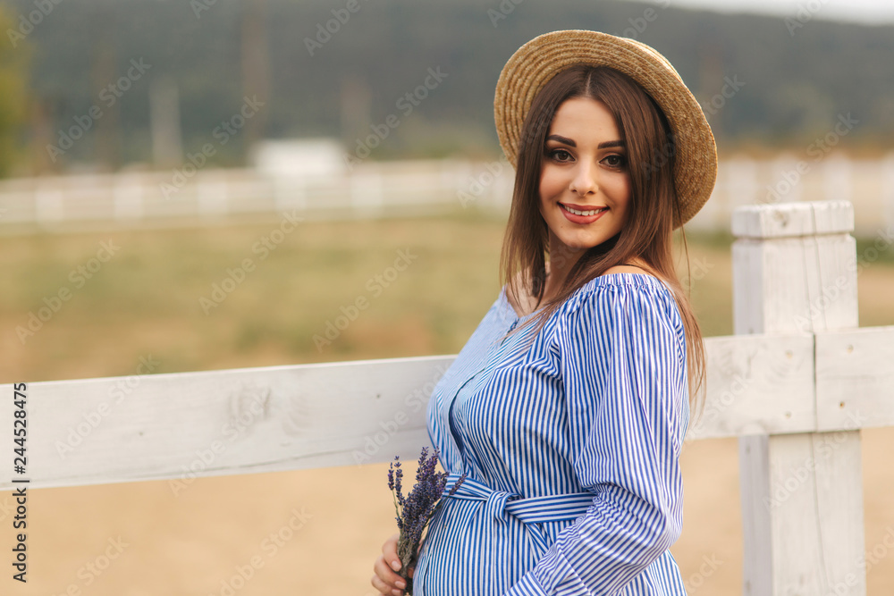 Beautiful pregnant woman in blue dress walking near the farm. Knitted bag and bouquet of lavender. Background of farm
