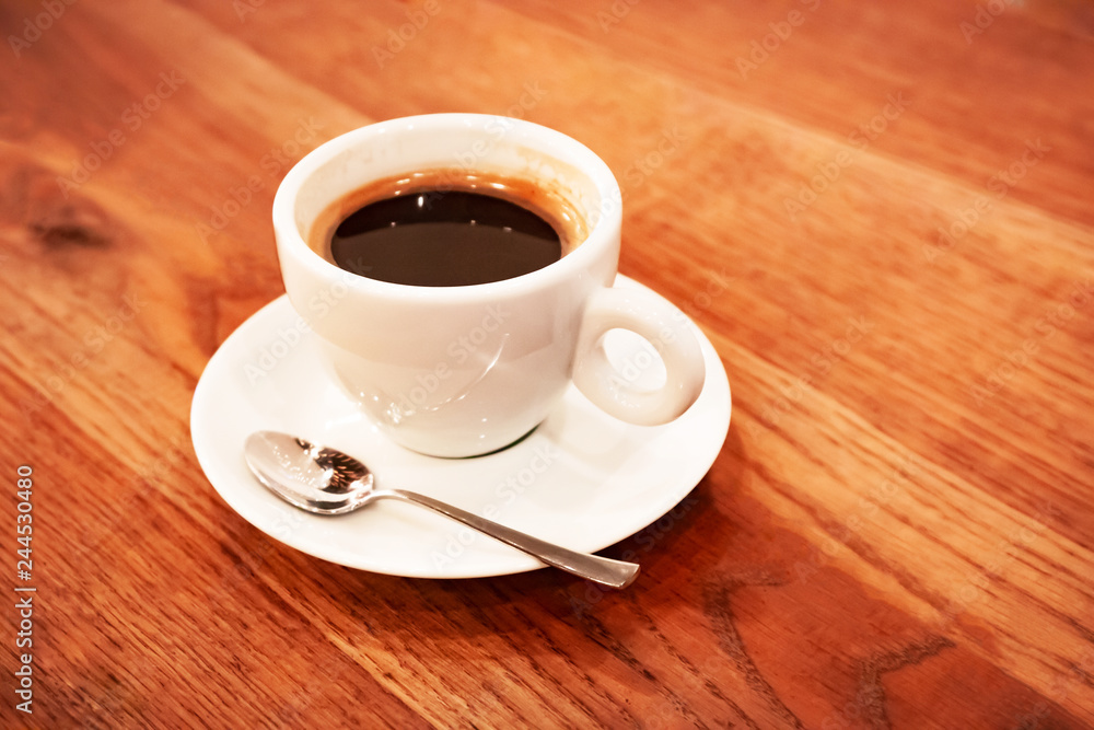 Close up white coffee cup with freshly brewed coffee and a spoon on a saucer stands on a dark wooden table.
