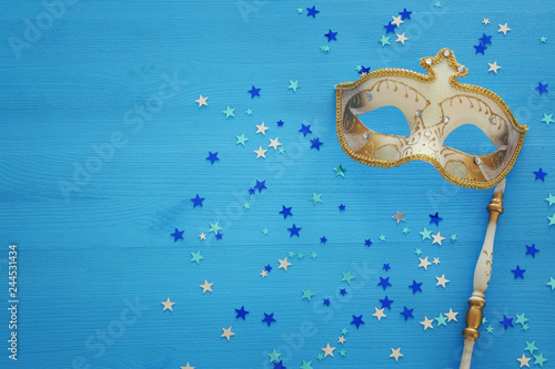 carnival party celebration concept with elegant gold mask on stick over blue wooden background and stars. Top view.