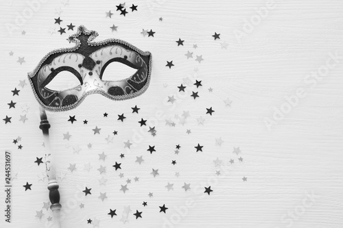 Black and white image carnival party celebration concept with elegant mask on stick over wooden background and stars. Top view.