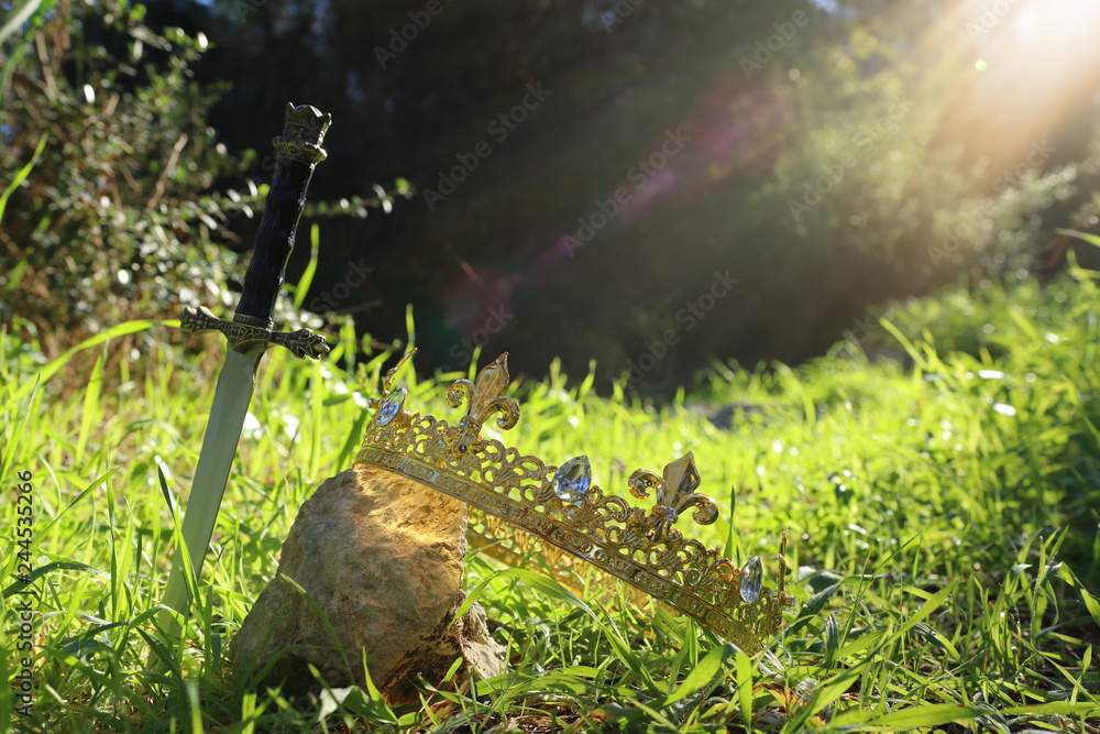 mysterious and magical photo of silver king crown and sword over the stone in the England woods or field landscape with light flare. Medieval period concept.