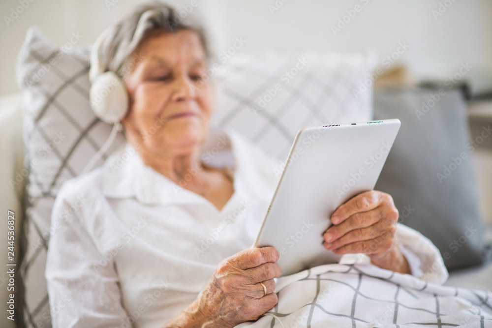 Sick senior woman with headphones and tablet lying in bed at home or in hospital. Sick senior woman with headphones and tablet lying in bed at home or in hospital.