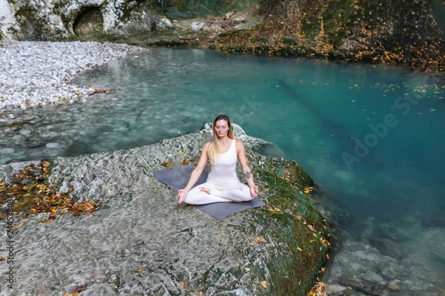 Fototapeta Naklejka Na Ścianę i Meble -  Top view - concentrated relaxed girl blonde yoga instructor meditates in lotus position with eyes closed while sitting on stone against backdrop of a picturesque mountain stream and green rock
