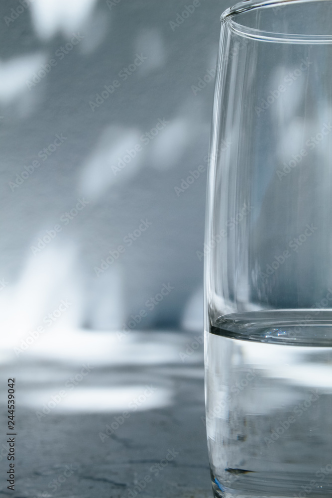 Close up of a glass of water standing in front of a white wall with shadows of leaves on a bright, sunny day