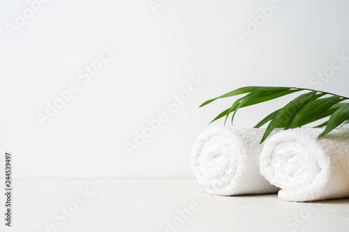 Spa concept: two white fluffy towels twisted into rolls on a light surface with a palm leaf with copy space