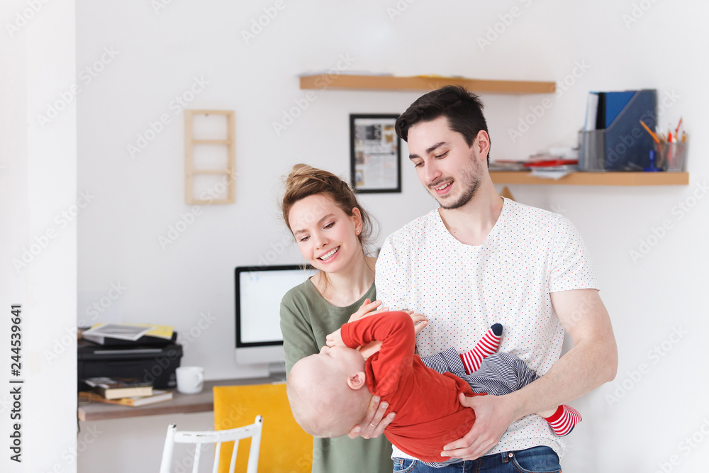Fototapeta premium Loving young couple with their one year old baby boy at home in white domestic interior. Beautiful happy family spend time together