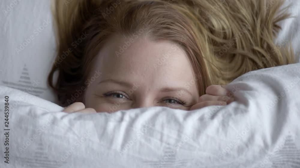 Closeup of Lovely young Woman Lies in Bed Covered with Blanket. Top view of Girl is Shy and Cover of her Face with Blanket. Female Smiling and Looking at Camera.