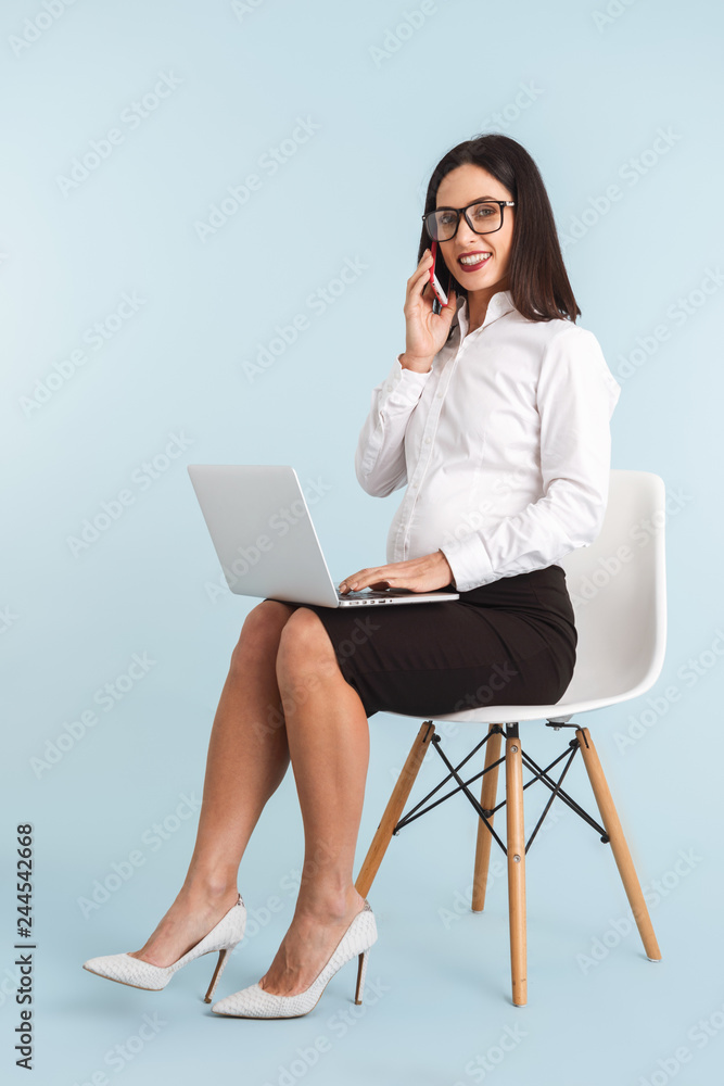 Pregnant business woman isolated over blue wall background using laptop computer talking by mobile phone. Pregnant business woman isolated over blue wall background using laptop computer talking by mobile phone.