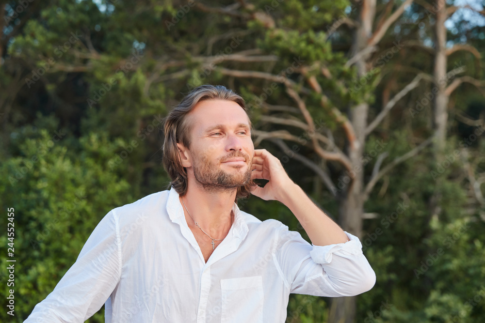 Portrait of the young handsome brutal man with long hair, the blue eyes, an easy bristle, he is dressed in a white shirt with a short sleeve, green trees on a background, light of a sunset Portrait of the young handsome brutal man with long hair, the blue eyes, an easy bristle, he is dressed in a white shirt with a short sleeve, green trees on a background, light of a sunset