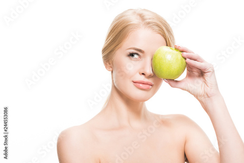 naked blonde girl holding green apple near eye and looking away isolated on white