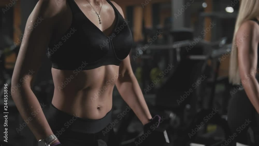 Close-up of gorgeous blonde fitness women exercising with dumbbells together at gym. Workout in dark gym in slow motion. Motivation healthy lifestyle. Fitness women in sport center Close-up of gorgeous blonde fitness women exercising with dumbbells together at gym. Workout in dark gym in slow motion. Motivation healthy lifestyle. Fitness women in sport center